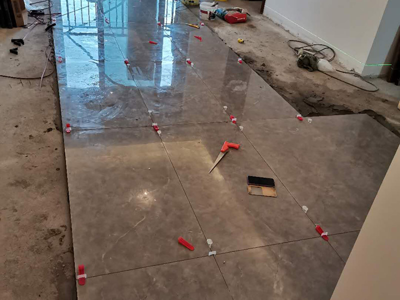 客廳地磚鋪貼：地磚鋪貼前，應(yīng)將地面清掃干凈，尤其是凝結(jié)后的混凝土，否則，地磚鋪貼后會(huì)出現(xiàn)翹邊、空鼓等現(xiàn)象，然后再灑水潤(rùn)濕地面。地面充分濕潤(rùn)，但不能有積水，然后將3:1混合好的沙子水泥鋪在地面上，進(jìn)行基層處理。在地磚背面均勻抹水泥砂漿作為粘合層，厚度不低于1cm，然后鋪在剛才試鋪的位置上，用橡皮錘輕敲地磚飾面直至密實(shí)平整達(dá)到要求。地磚鋪完后24h進(jìn)行清理勾縫，勾縫前應(yīng)先將地磚縫隙內(nèi)雜質(zhì)擦凈，用專(zhuān)用填縫劑勾縫，完工后（一般宜在24h之后）再用棉紗等物對(duì)地磚表面進(jìn)行清理。